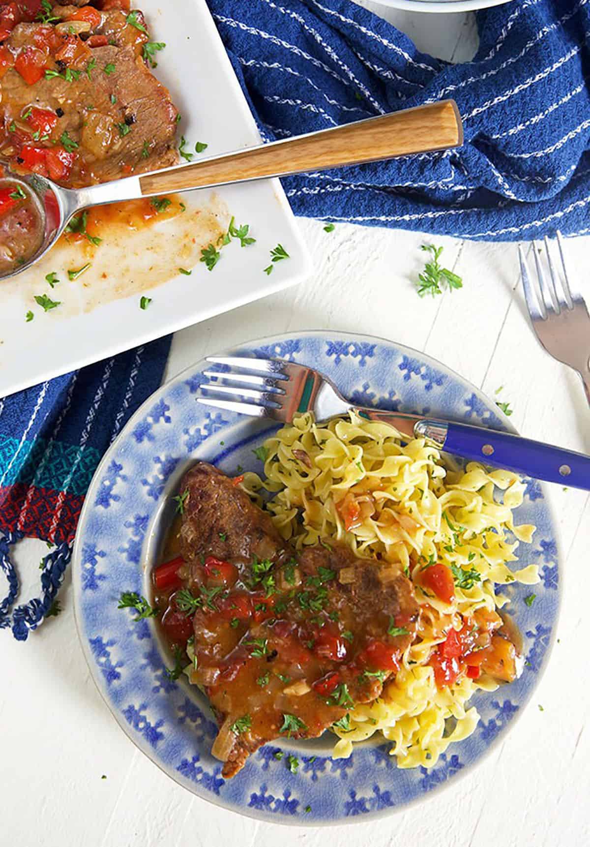 Swiss steak shown on a blue plate with egg noodles and a fork.