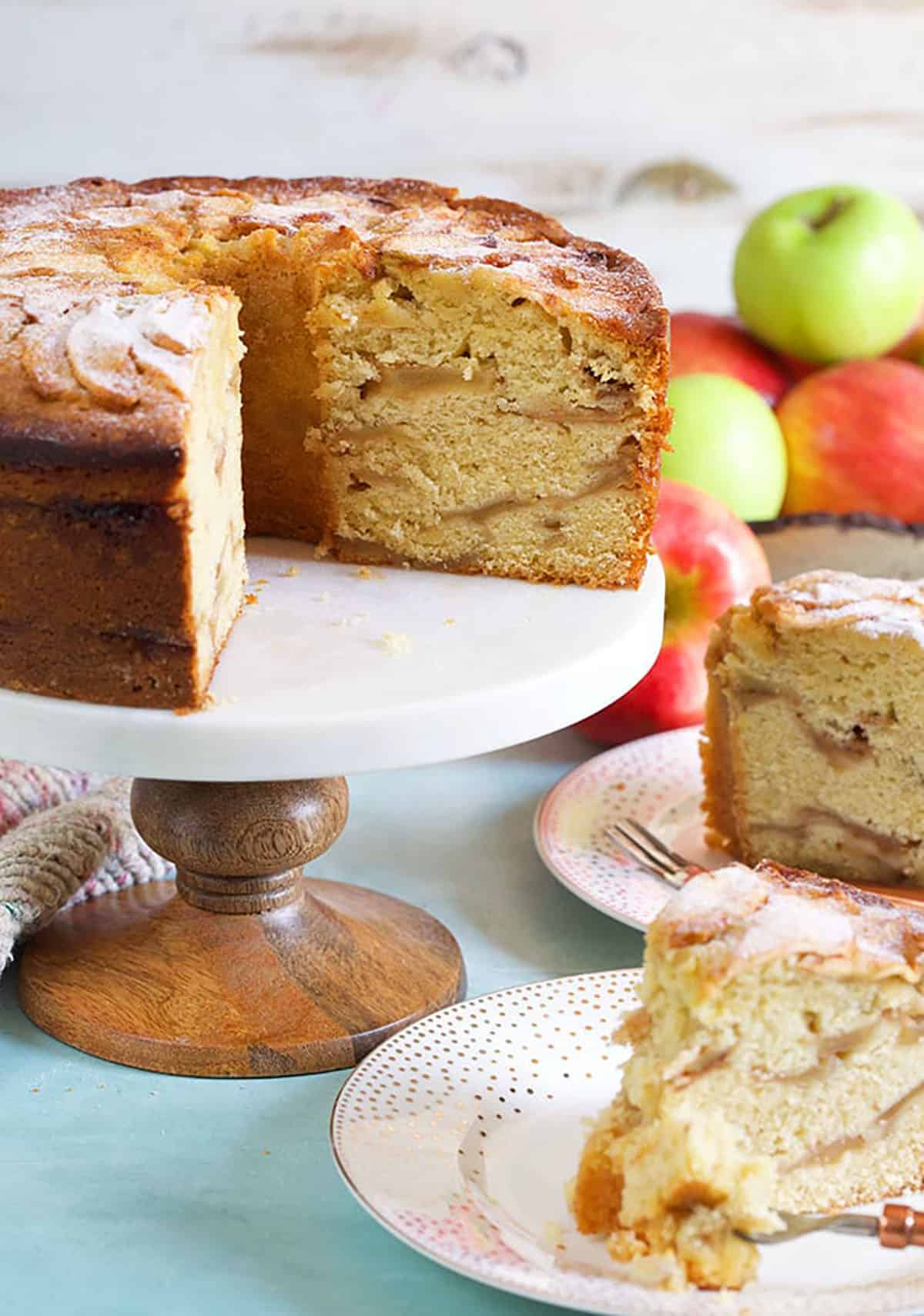Jewish apple cake with two slices cut out and apples in the background.