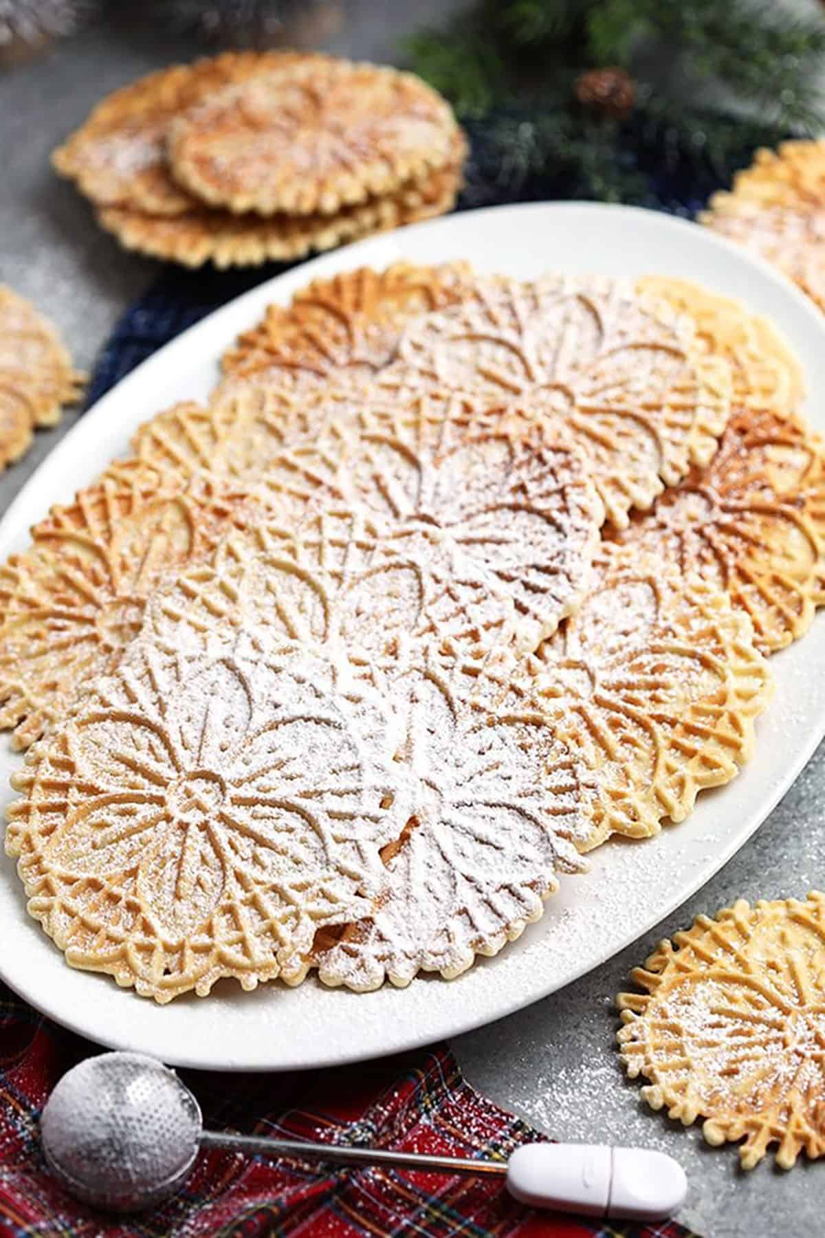 Platter of pizzelles on a white platter with a sugar wand on a plaid napkin.