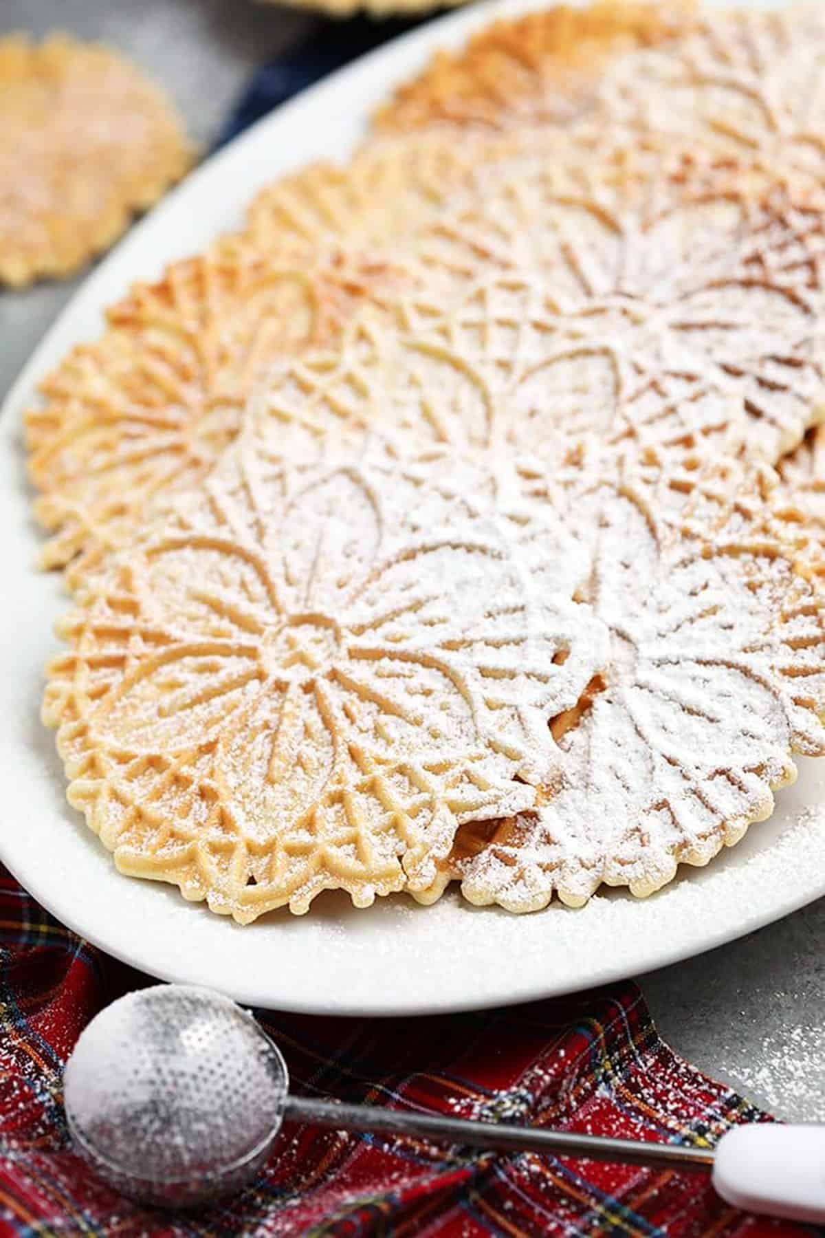 Homemade Pizzelles on a white platter with a plaid napkin underneath.