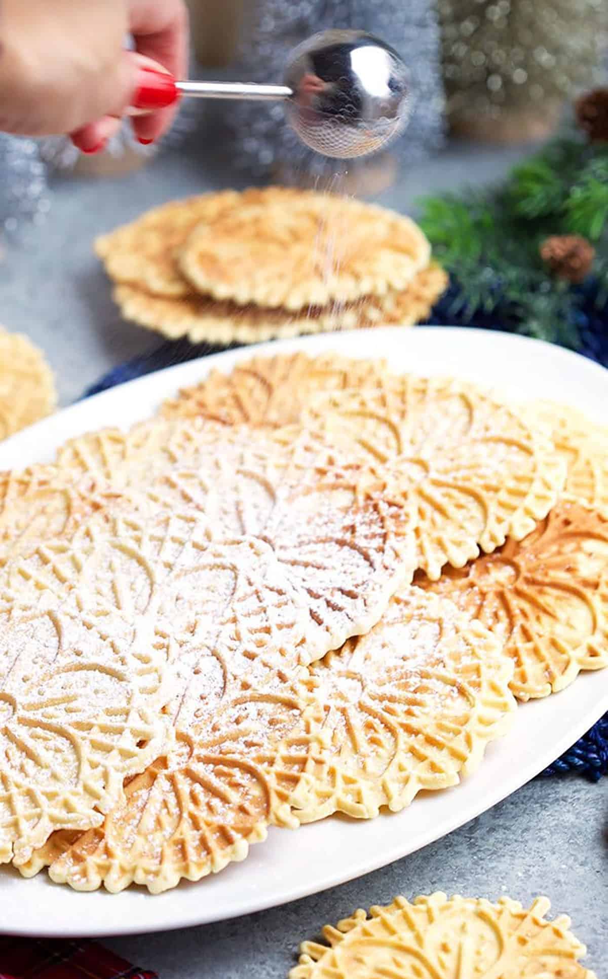 Powdered sugar wand sprinkling sugar over a platter of homemade pizzelles on a white platter.