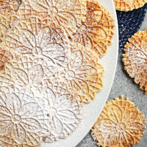 Overhead shot of pizzelles on a white platter with powdered sugar and a plaid napkin.