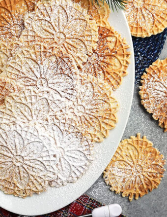 Overhead shot of pizzelles on a white platter with powdered sugar and a plaid napkin.