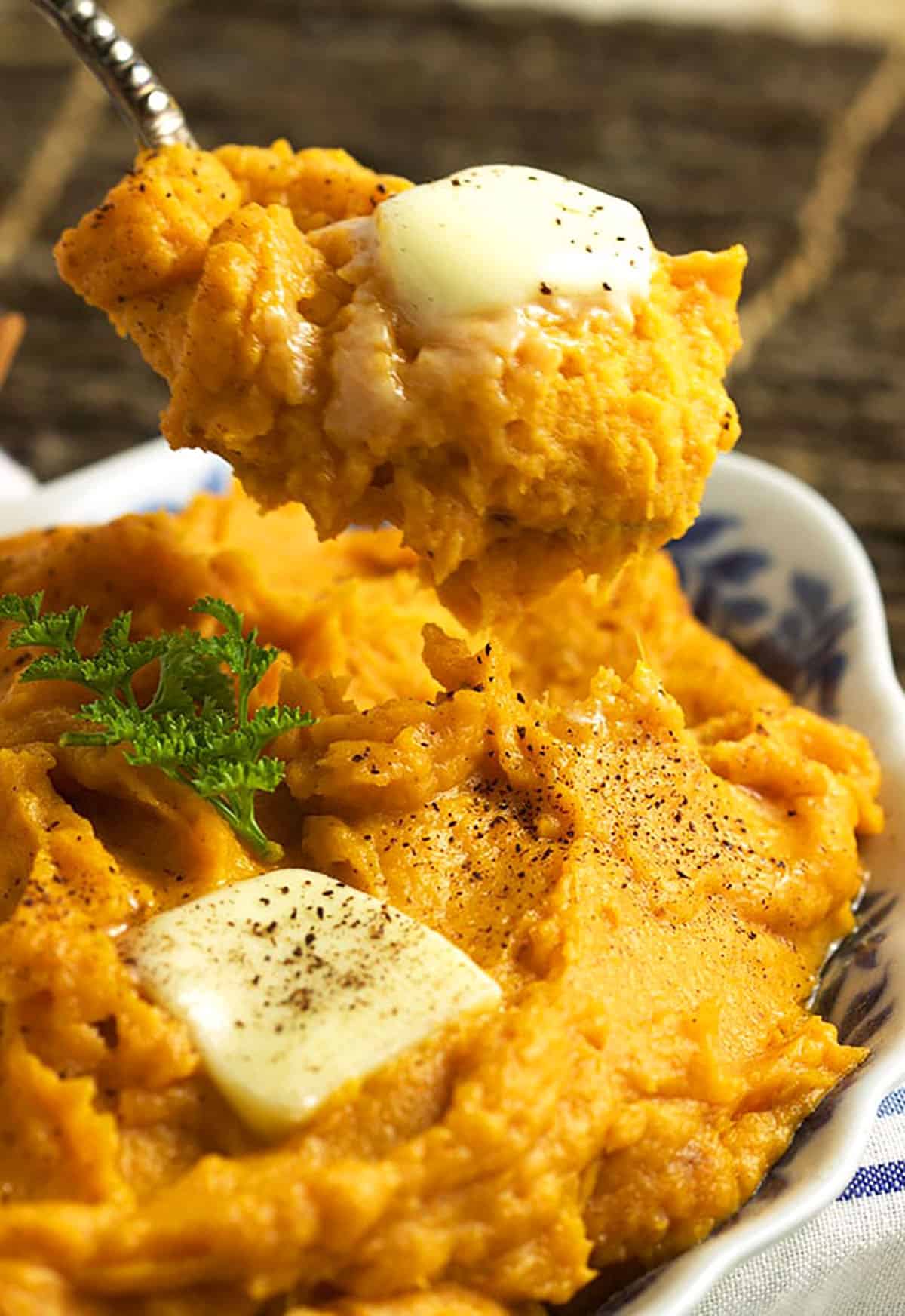 Serving spoon with mashed sweet potatoes on it topped with a pat of butter over a bowl of sweet potatoes.
