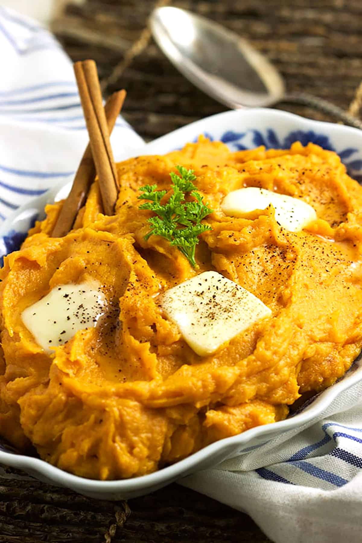 Mashed Sweet potatoes in an oval blue and white bowl topped with three pats of butter and a sprig of parsley.