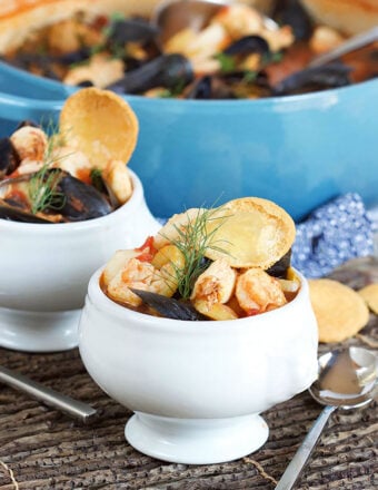 Bouillabaisse in a white footed bowl with a blue pot in the background.