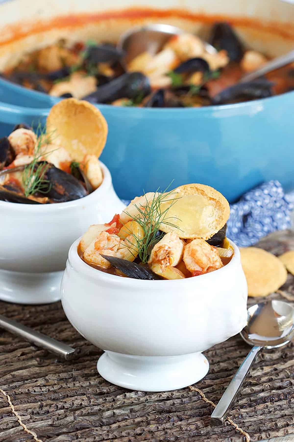 Bouillabaisse in a white footed bowl with a blue pot in the background.