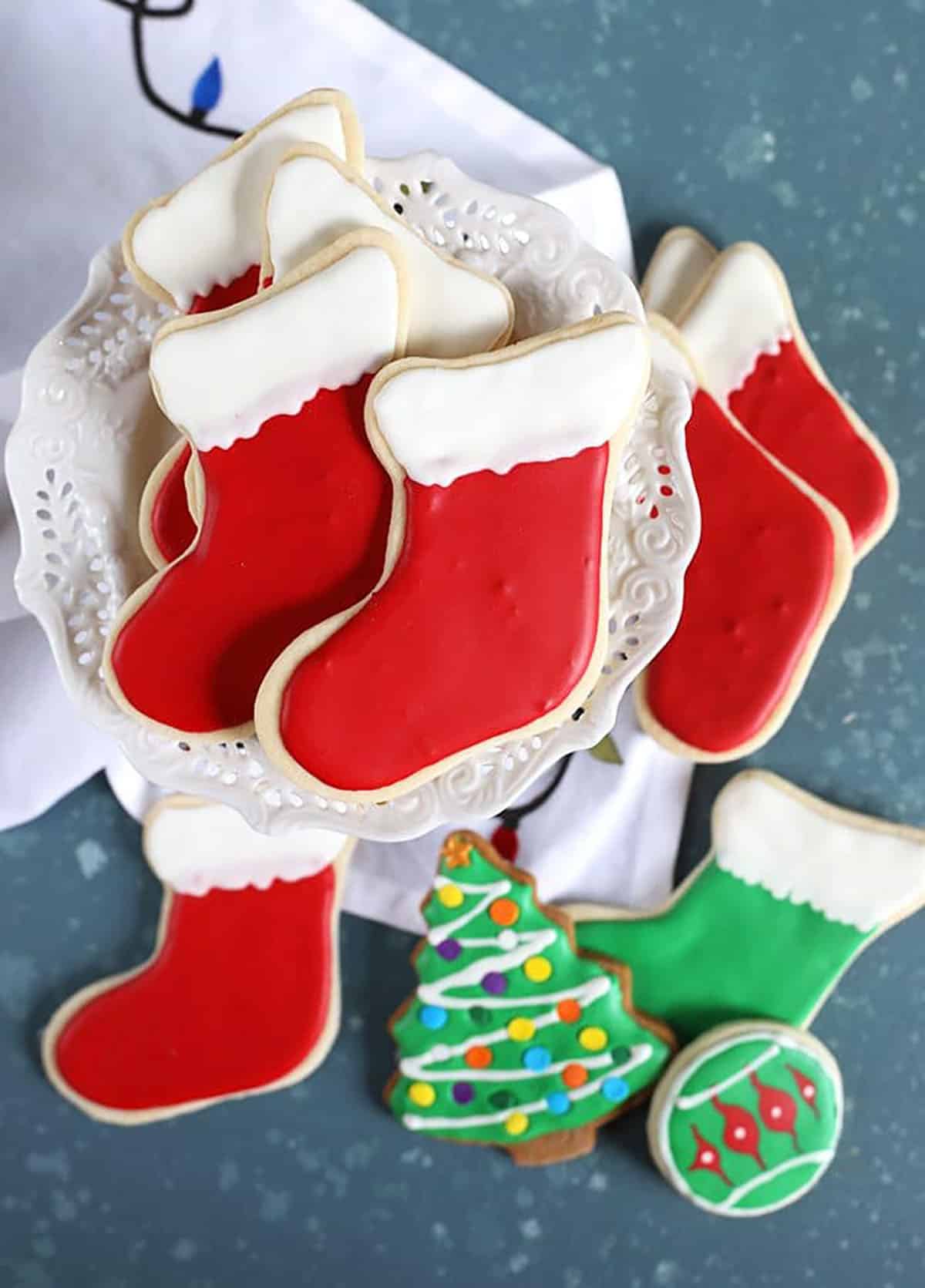 Stocking cut out cookies on a cake plate with a christmas tree cookie on a blue background.