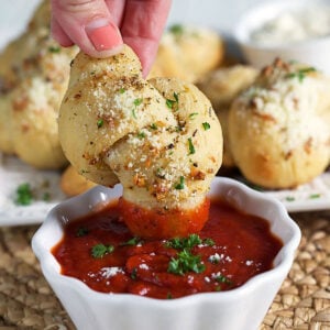 A homemade garlic knot is dipped into a small bowl of red sauce.