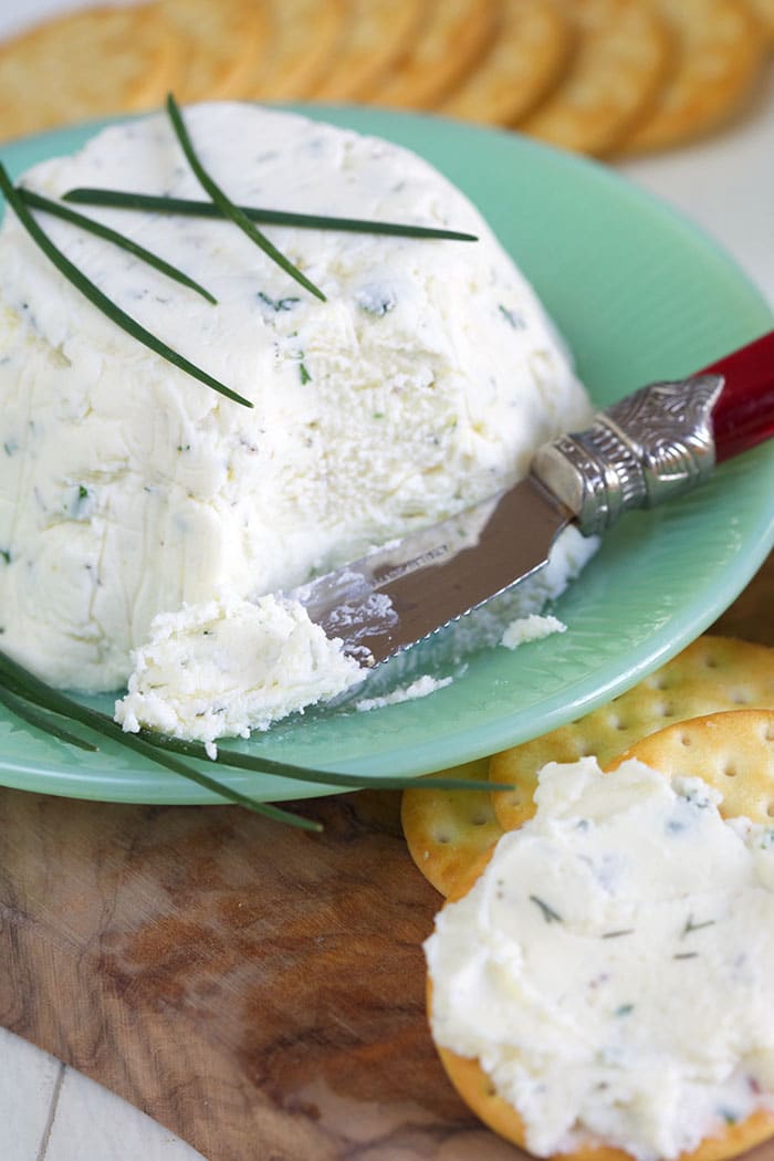 A cheese knife has cut into the white cheese and is smearing it onto a cracker.
