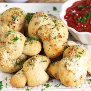 A number of garlic knots sit on a white plate, next to a small white bowl of red sauce.