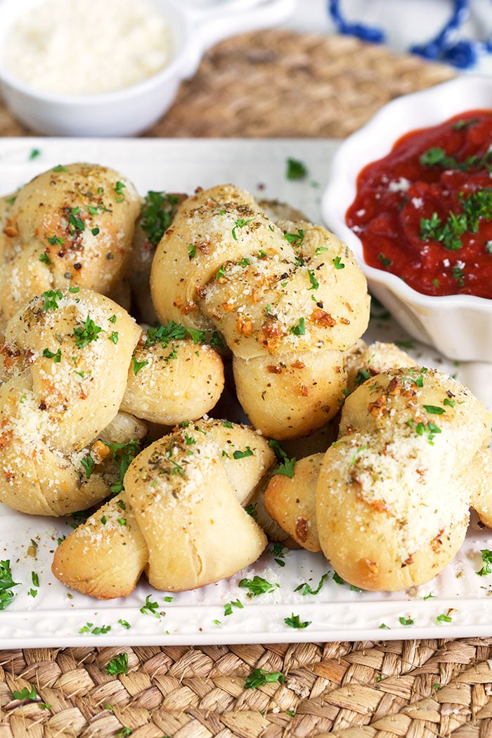 A number of garlic knots sit on a white plate, next to a small white bowl of red sauce.