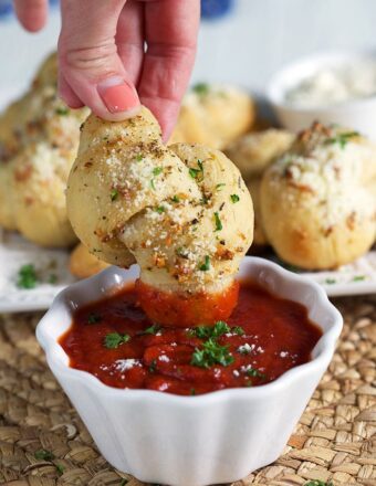 A homemade garlic knot is dipped into a small bowl of red sauce.