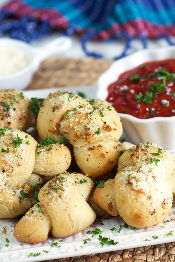 A stack of garlic knots is on a white plate, next to a small red bowl of garlic sauce.