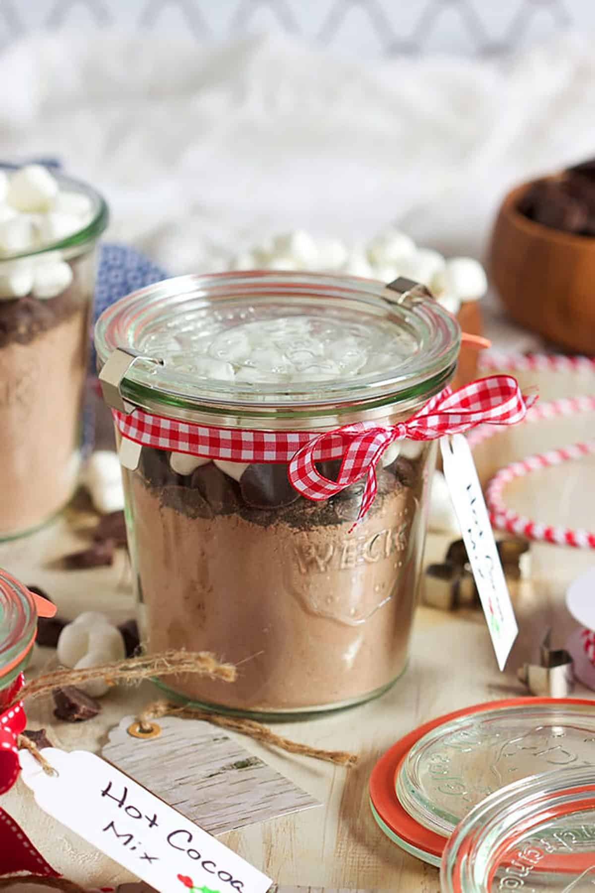 glass jar filled with hot cocoa mix chocolate chunks and marshmallows with a glass lid and a red checked ribbon.