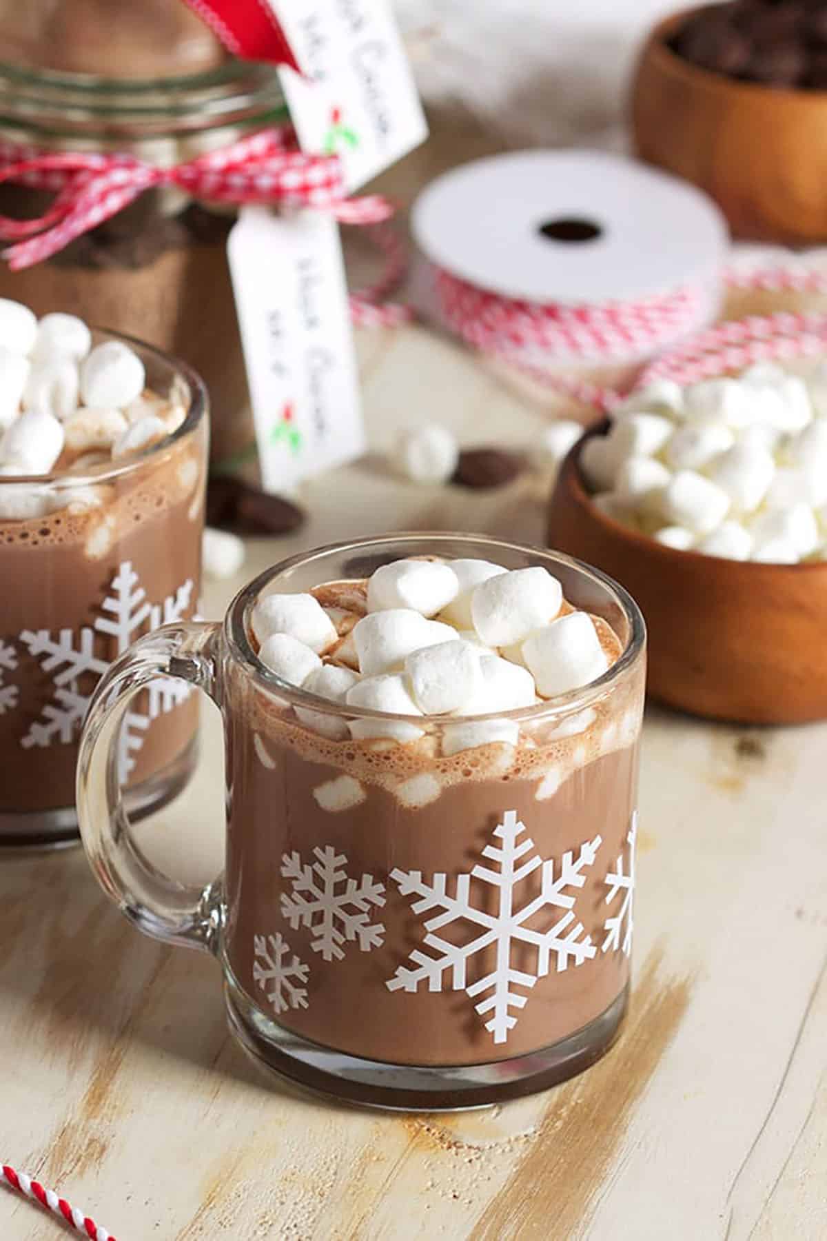 Hot cocoa in a glass mug with snowflakes on it and jars of hot cocoa mix in the background.