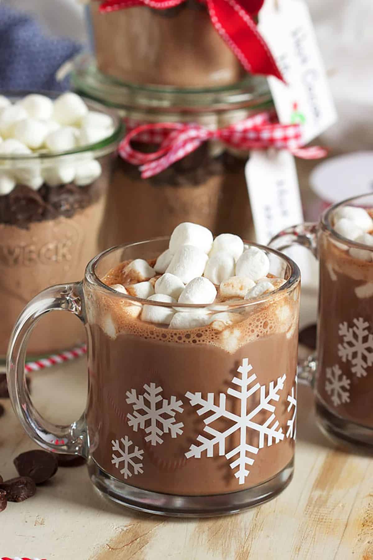 Hot chocolate with marshmallows on top in a glass mug on a wood background with a jar of cocoa mix in the background.