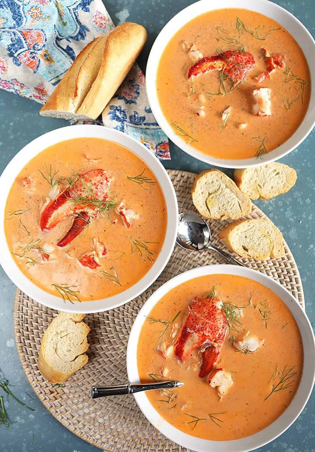 Overhead shot of three white bowls filled with lobster bisque with lobster claws in the center of each bowl and a broken baguette on the side.