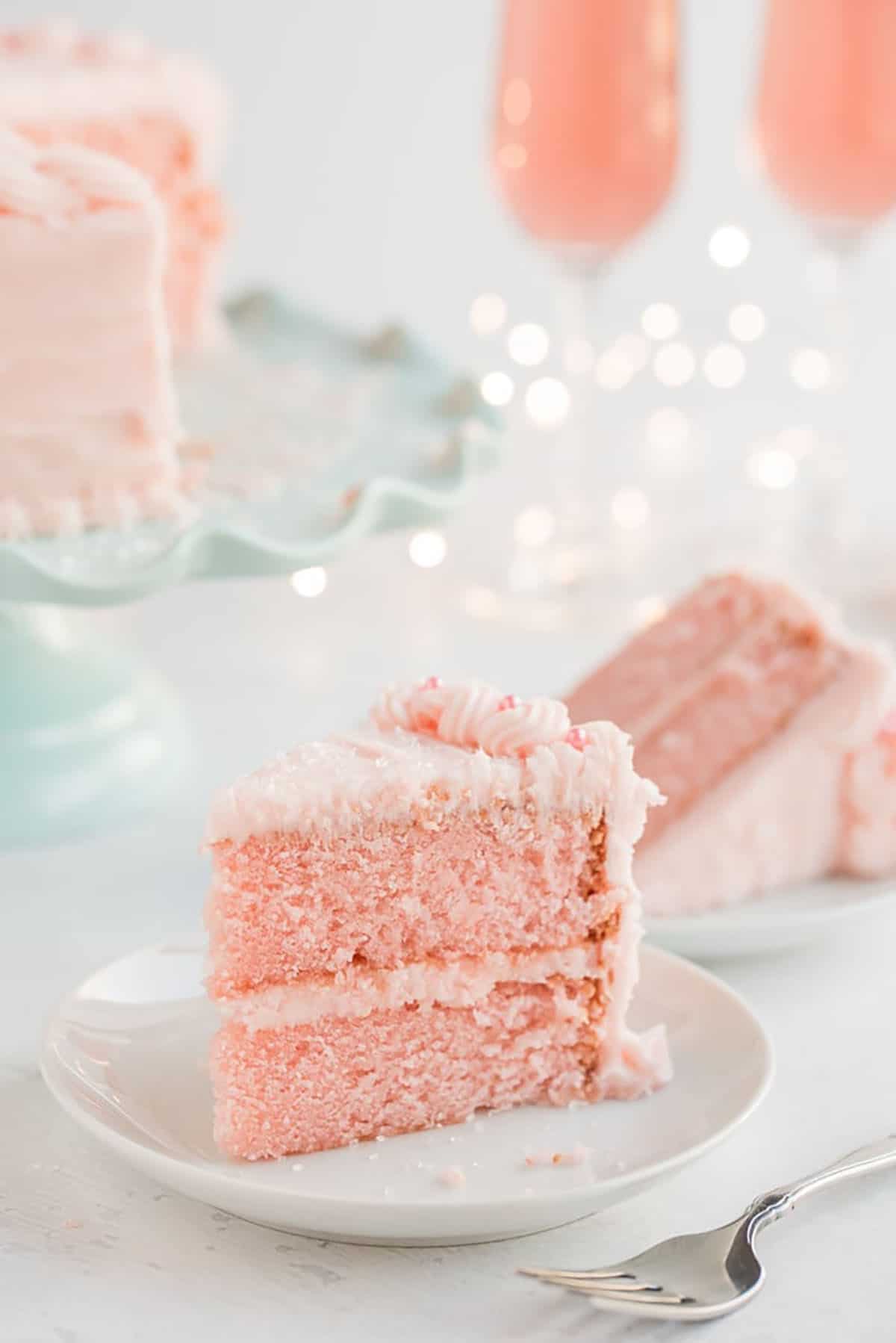 Pink Champagne Cake slice on a white plate with glasses of champagne in the background.