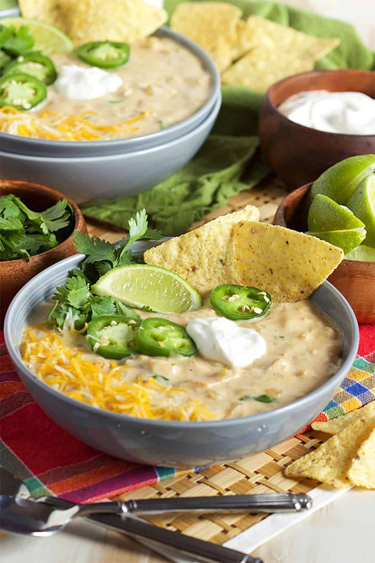 White chicken chili in a gray bowl topped with sliced jalapenos, a lime wedge, shredded cheese and sour cream.