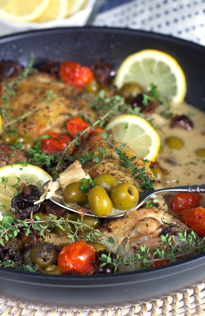 A spoon is holding several green olives above a skillet full of chicken provencal.