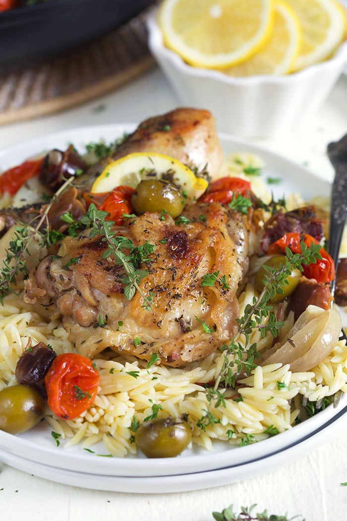 Roasted chicken, olives and herbs sit atop a serving of rice on a white plate.