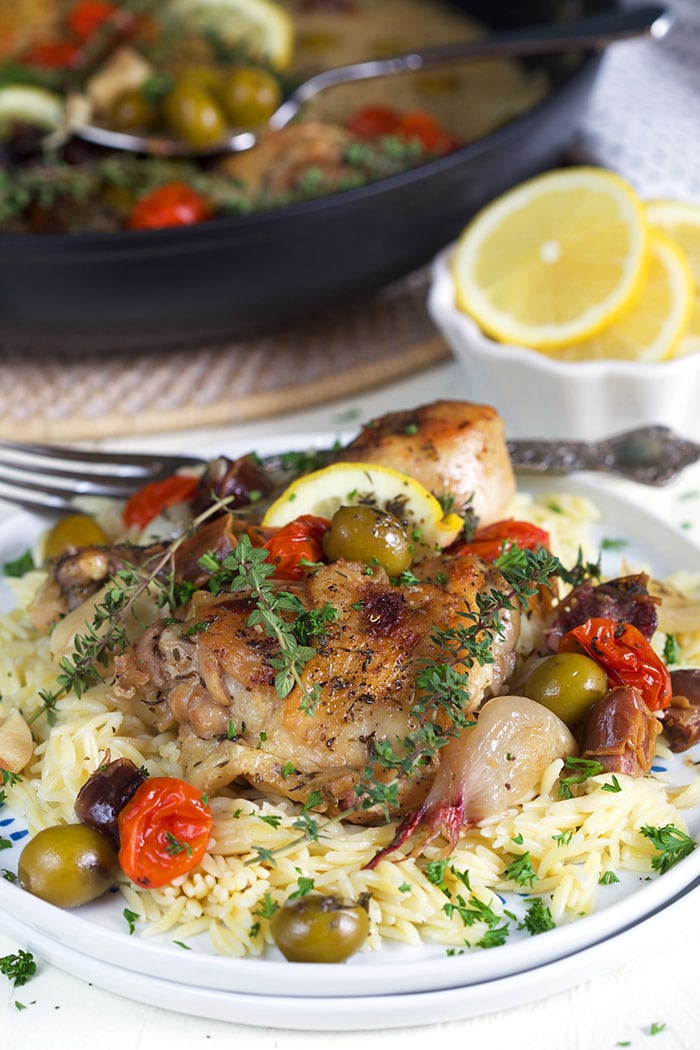 A plate of chicken provencal and rice is placed next to a black skillet and white bowl of lemon slices.