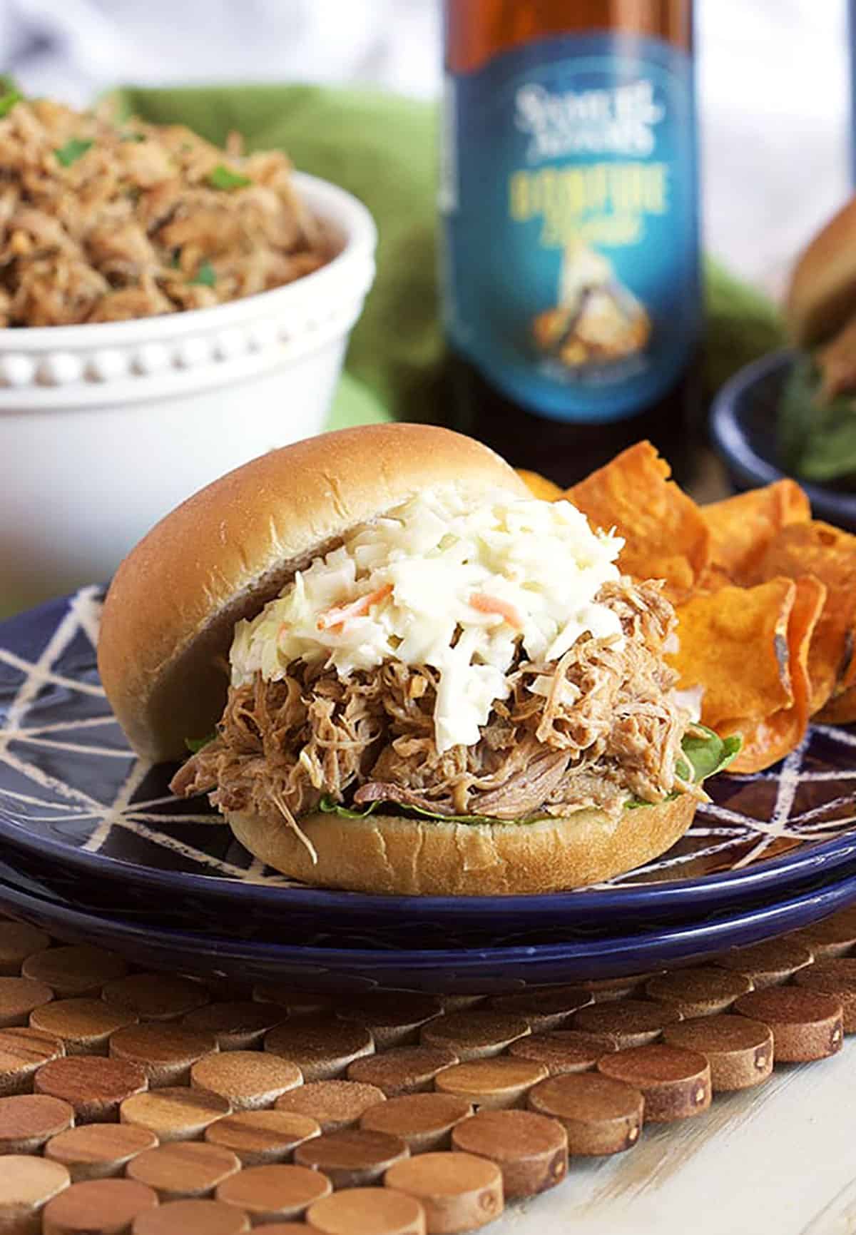 Slow cooker pulled pork sandwich with coleslaw on top on a blue plate with sweet potato chips and a bottle of beer in the background.