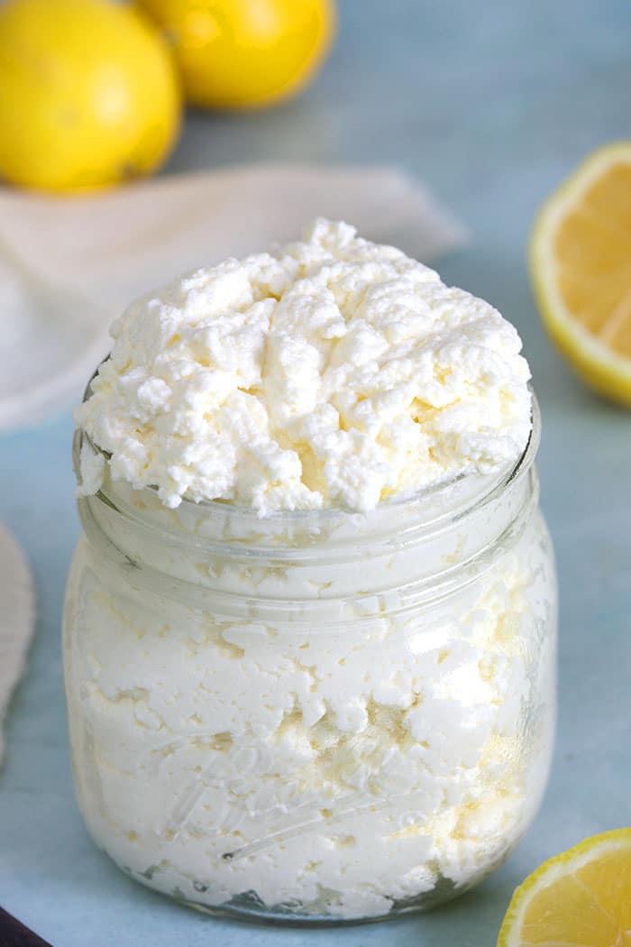A glass jar of ricotta cheese is on a counter with sliced lemons.