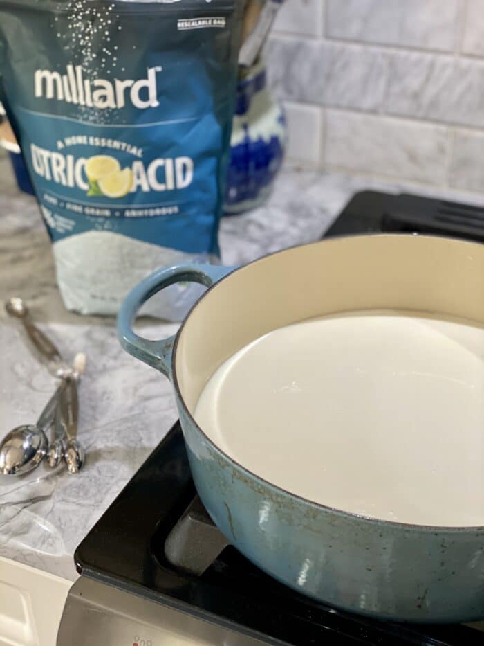 A large blue pot is heating milk on an oven, next to a bag of citric acid.