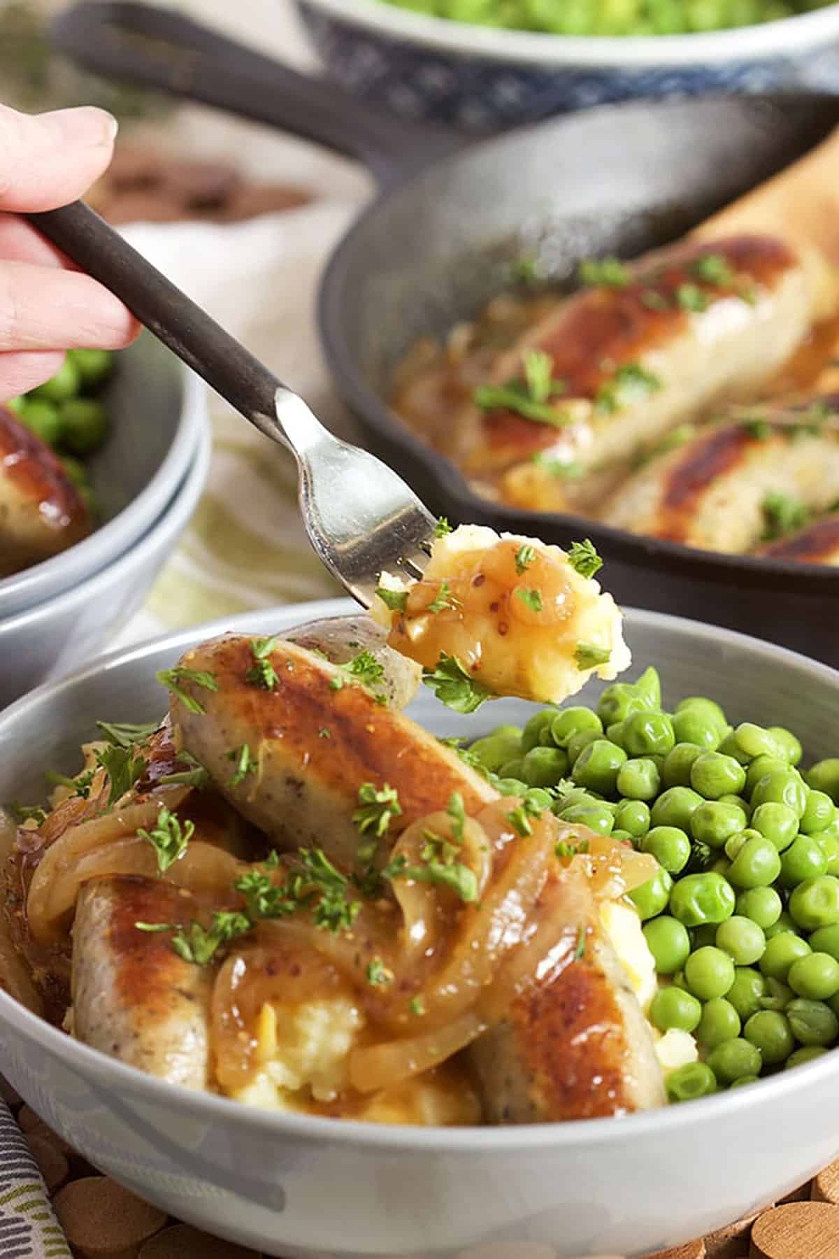Bangers and Mash in a gray bowl with green peas and a fork with mashed potatoes on it.