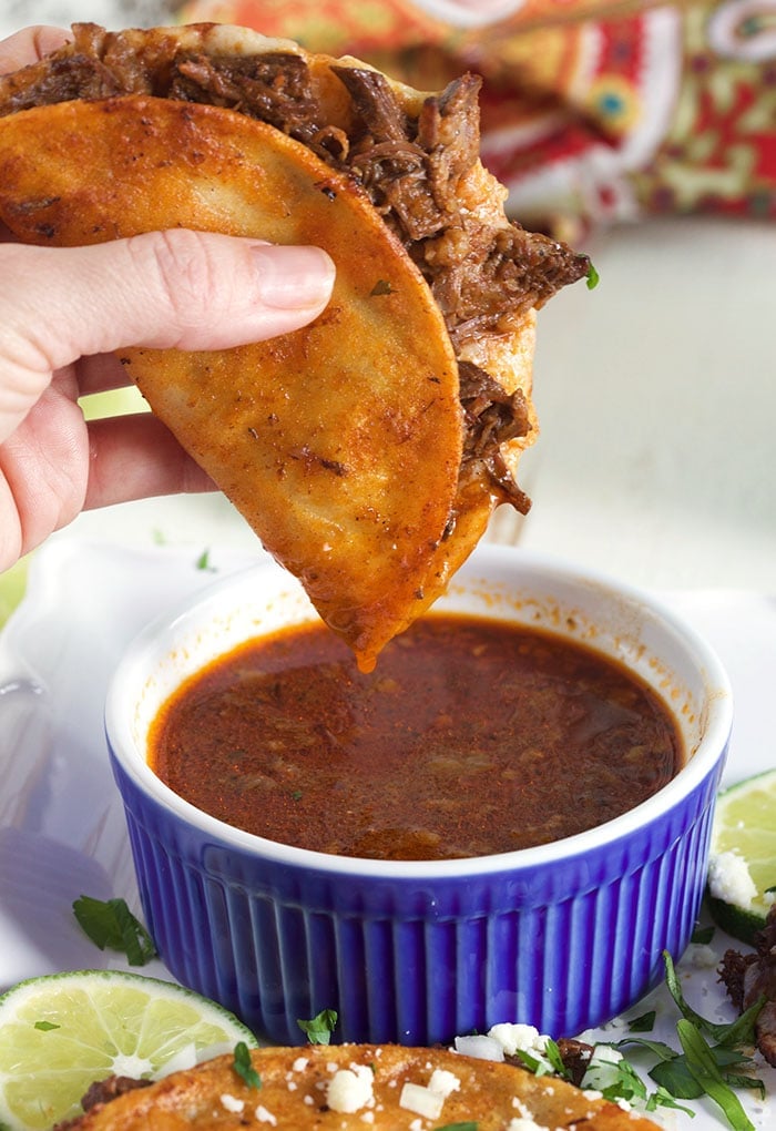 A crispy taco is being held above a small bowl of cooked stew.
