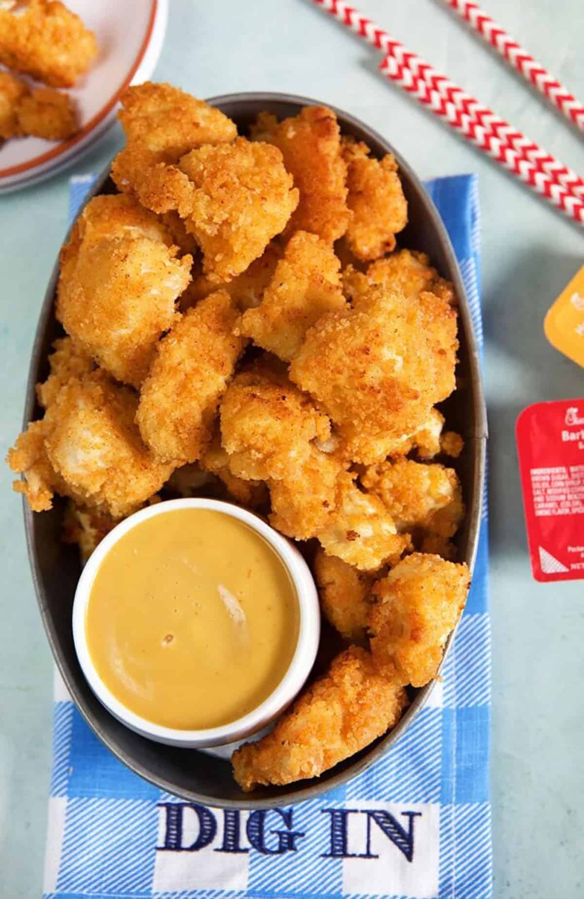 Steel tray filled with chicken nuggets with Chick Fil A sauce in a white dish on a blue and whit checkered napkin that says Dig In.