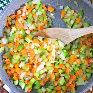 A wooden spoon is mixing a fully cooked mirepoix in a large skillet.