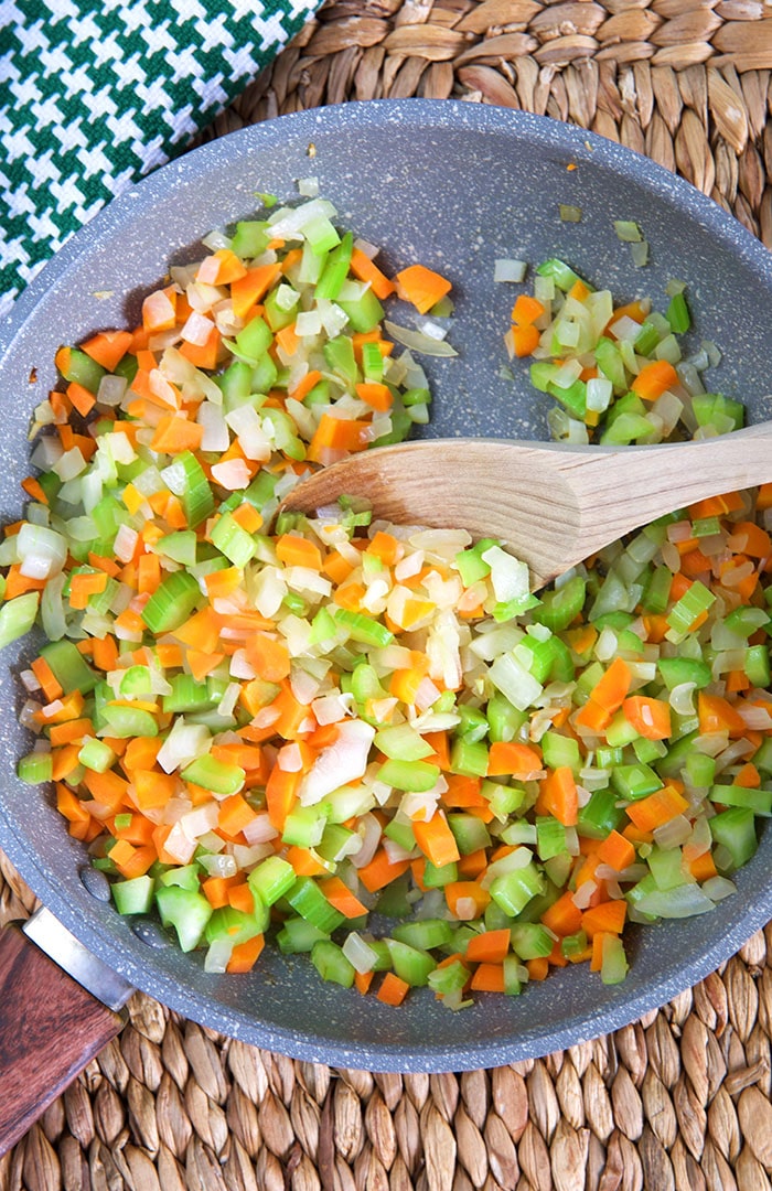 A wooden spoon is mixing a fully cooked mirepoix in a large skillet. 