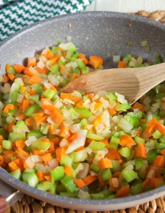 Onions, celery and carrots are being stirred in a large skillet with a wooden spoon.