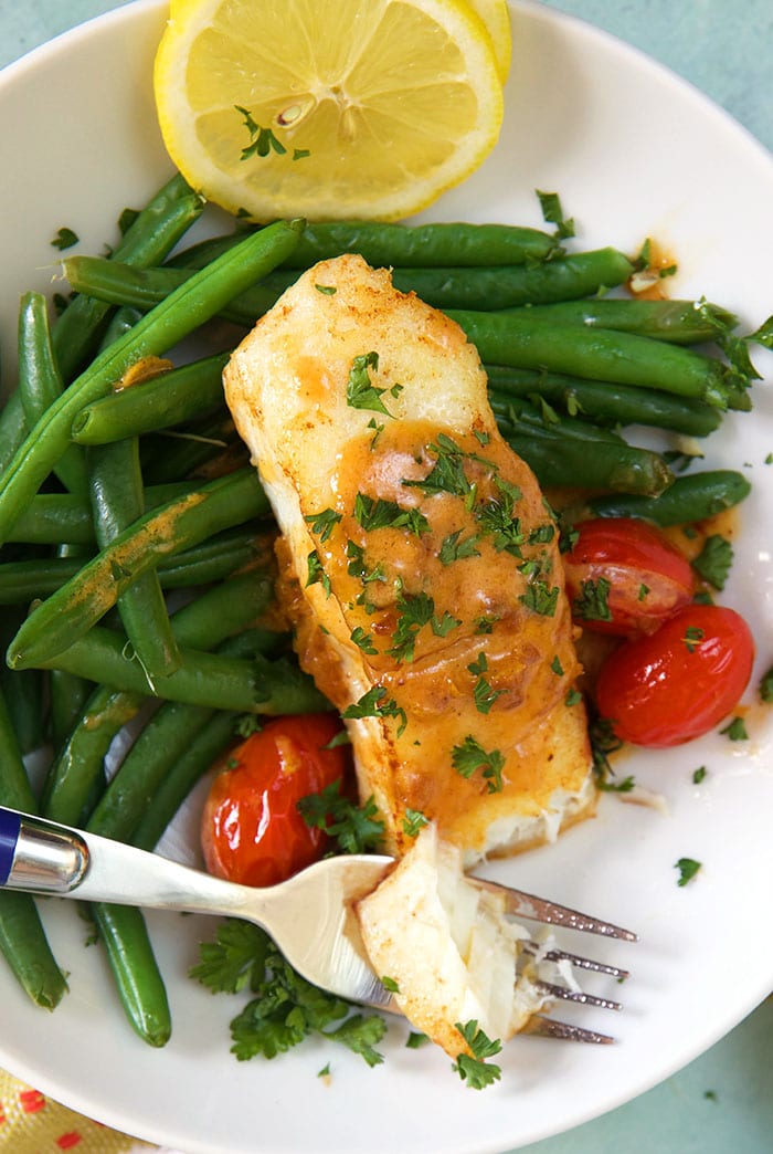 A piece of cooked halibut sits on top of green beans and tomatoes on a white plate.