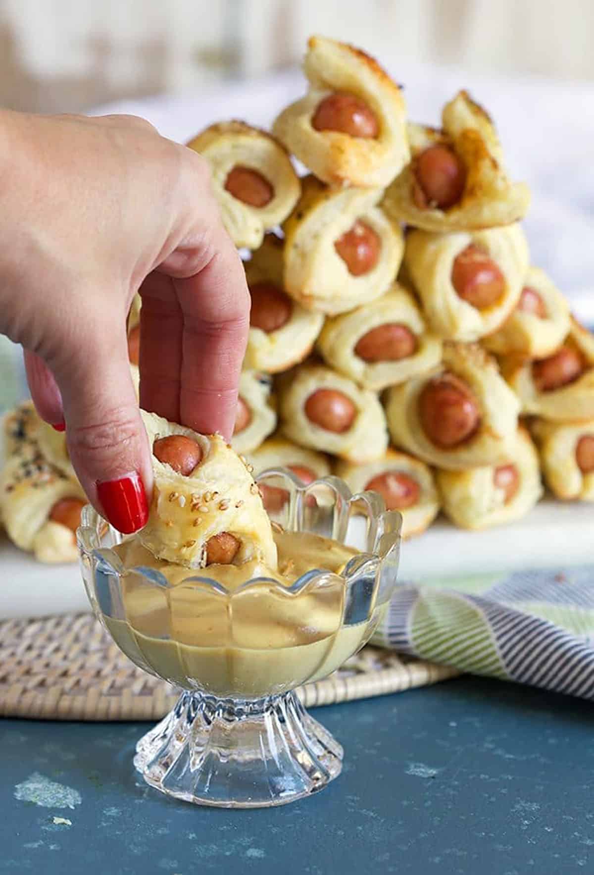 Pigs in a Blanket being dipped into mustard in a glass dish on a blue background.