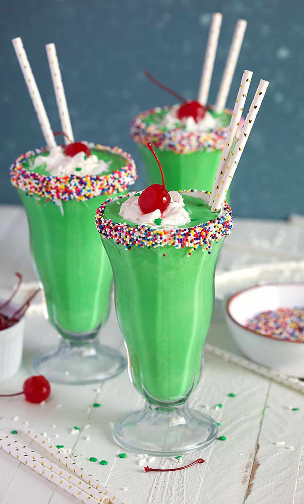 Three shamrock shake milkshakes in glasses with paper straws on a white board with a blue background.