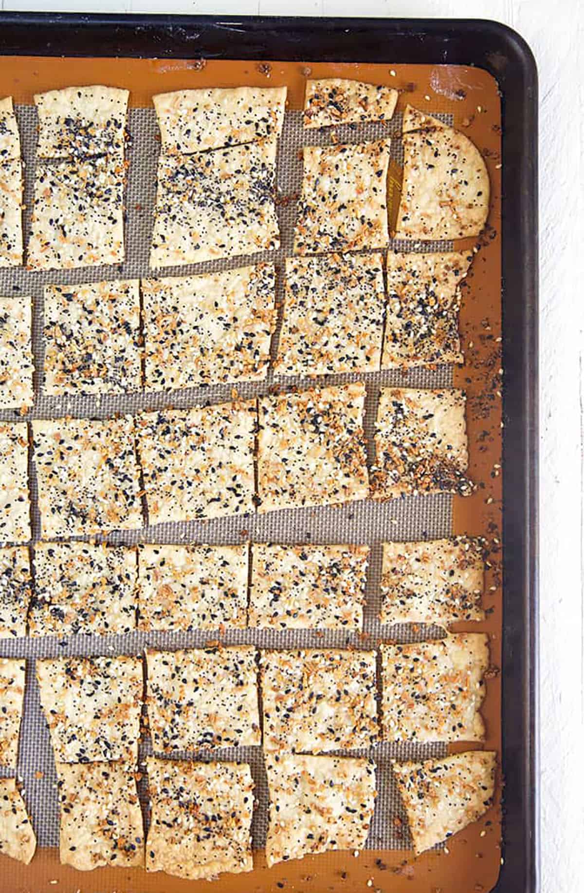 Many pieces of everything crackers are spread out on a large lined baking sheet.