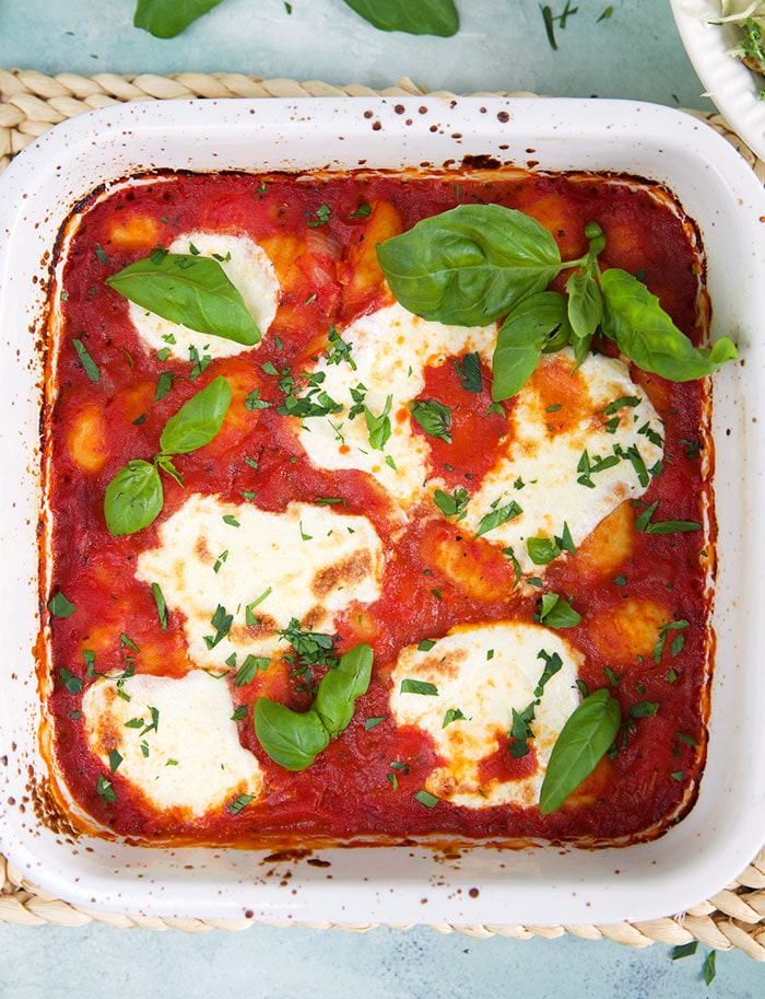 A square baking pan contains red sauce, gnocchi, white cheese and fresh basil.