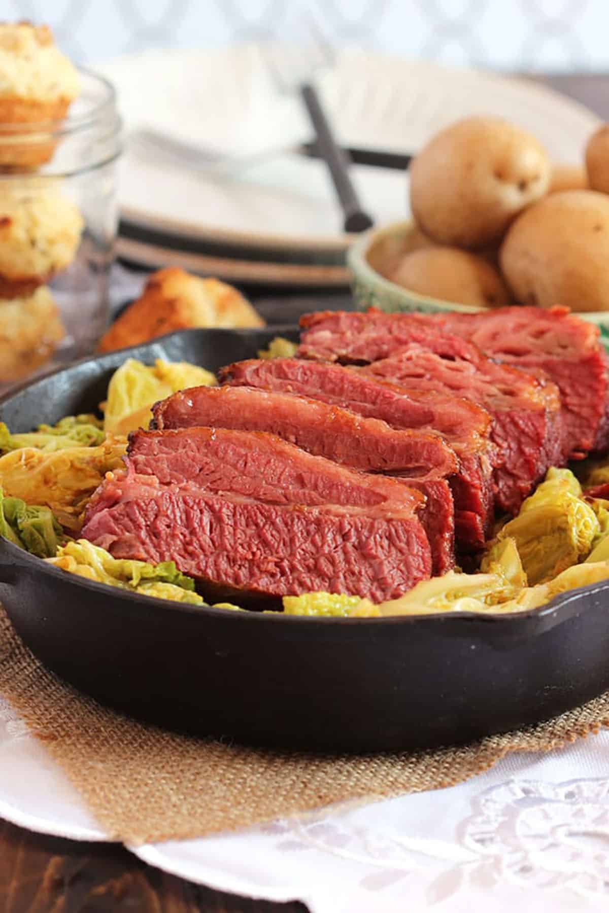 sliced corned beef on a bed of cabbage in a cast iron skillet with muffins in the background.