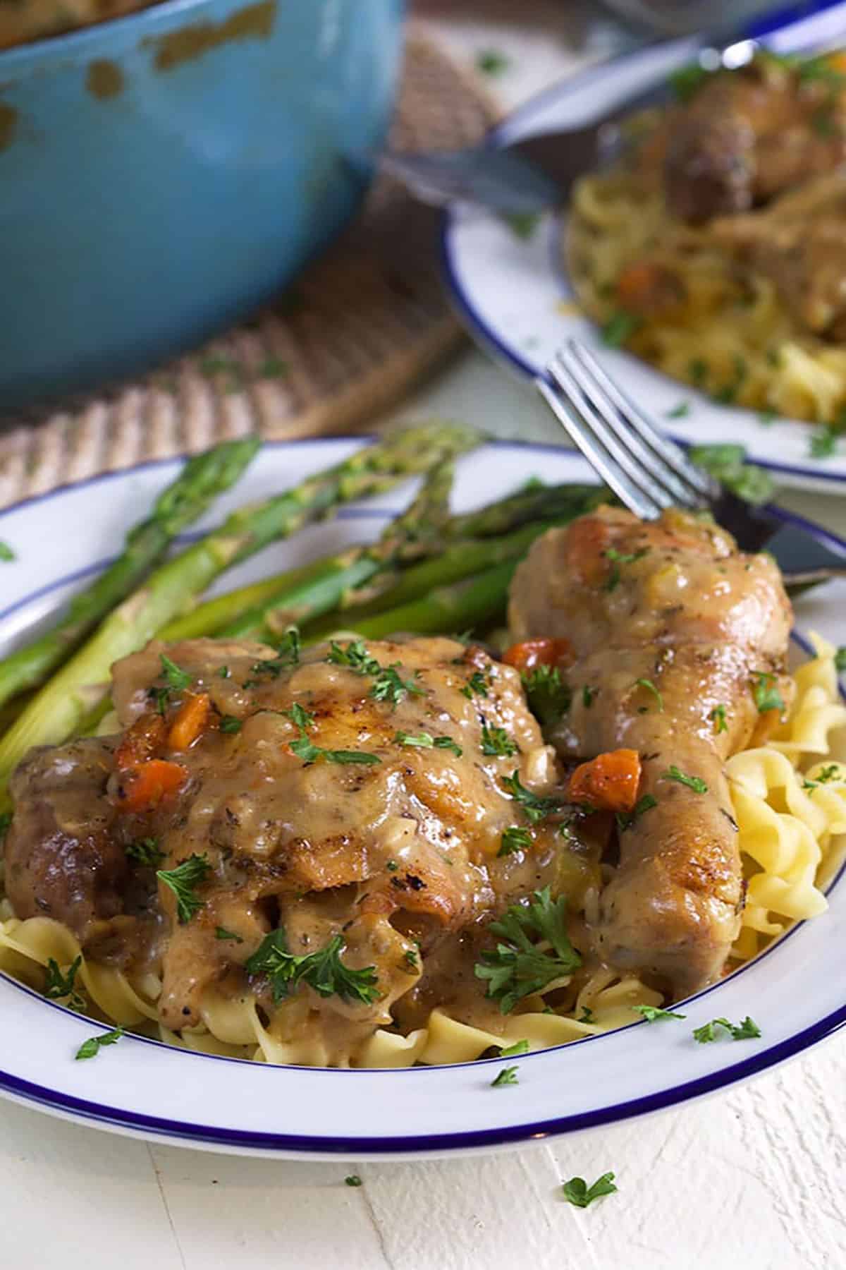 A serving of chicken fricassee is served over cooked egg noodles and asparagus on a white plate.