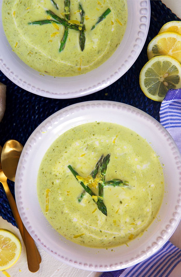 Two bowls of creamy asparagus soup are placed next to lemon slices.