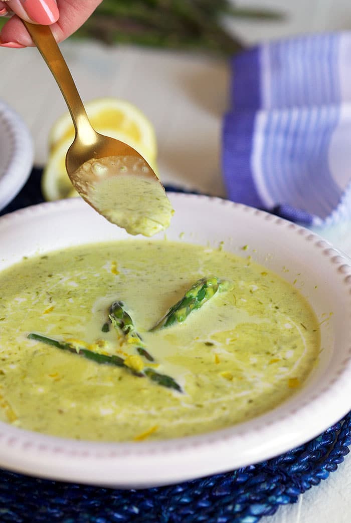 A spoon is lifting a bite sized portion of creamy asparagus soup out of a white bowl.