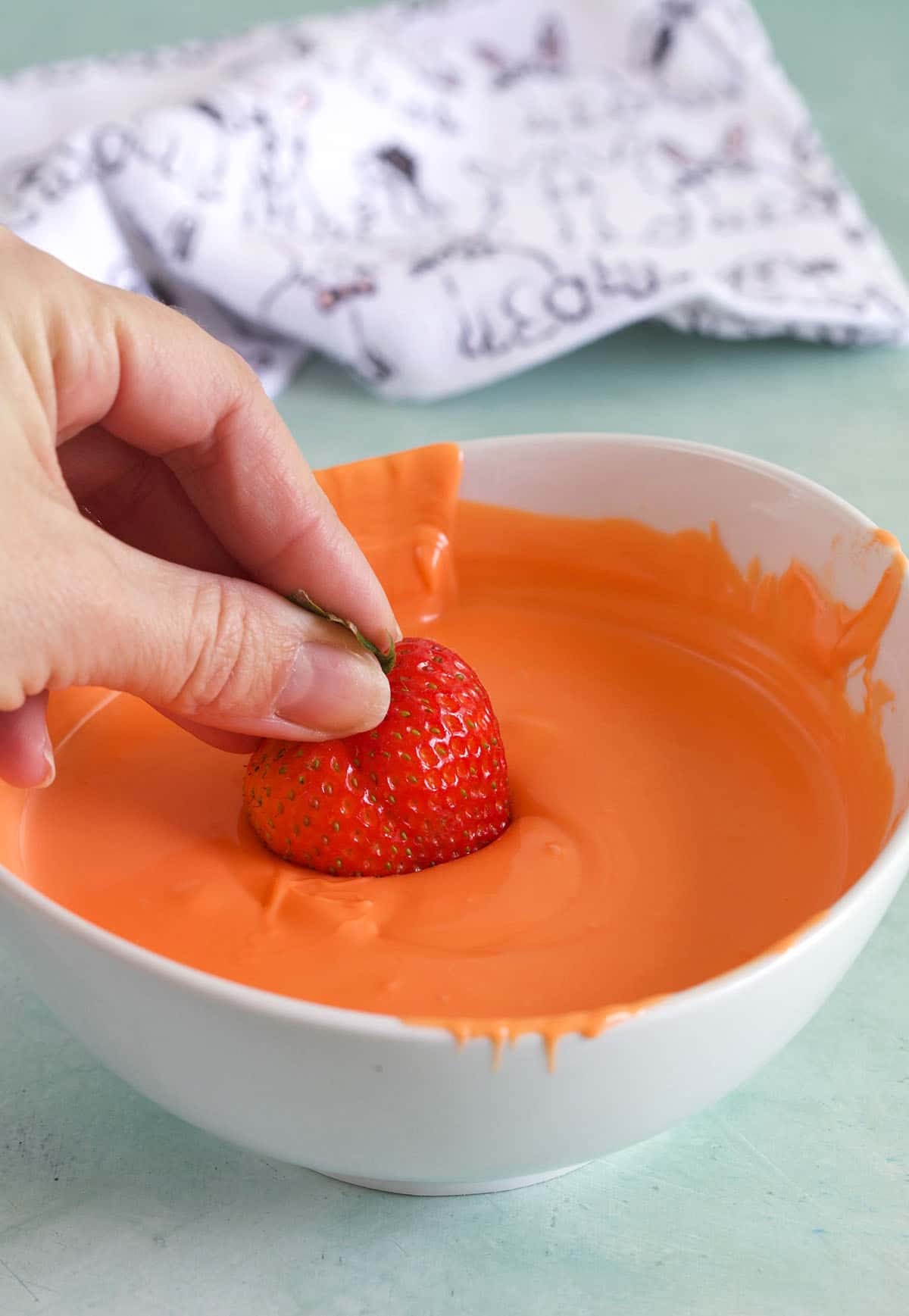 A strawberry is being dipped into a bowl of melted white chocolate that's been dyed orange.