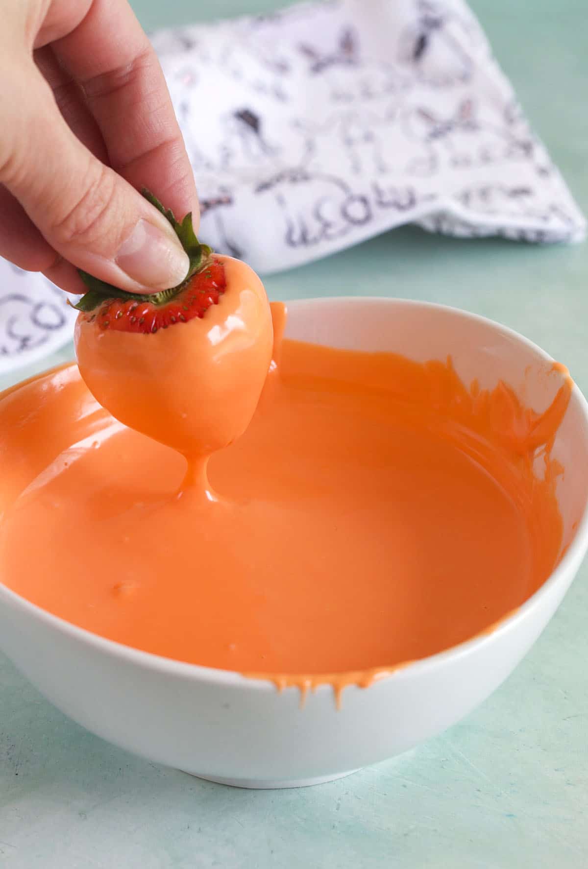 A strawberry dipped in orange chocolate is being removed from the white bowl. 