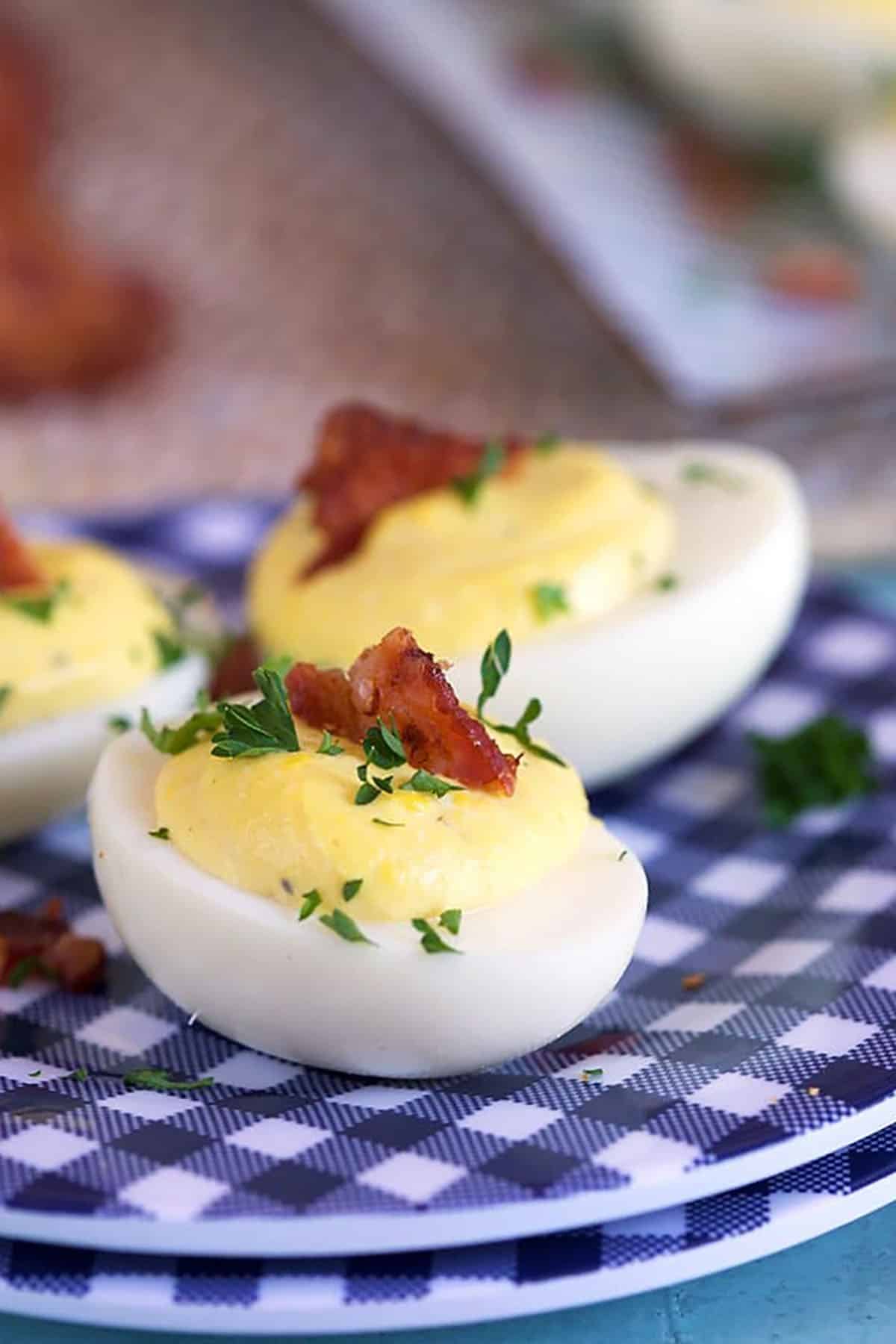 Creamy deviled eggs on a blue and white gingham plate with a slice of bacon on top.