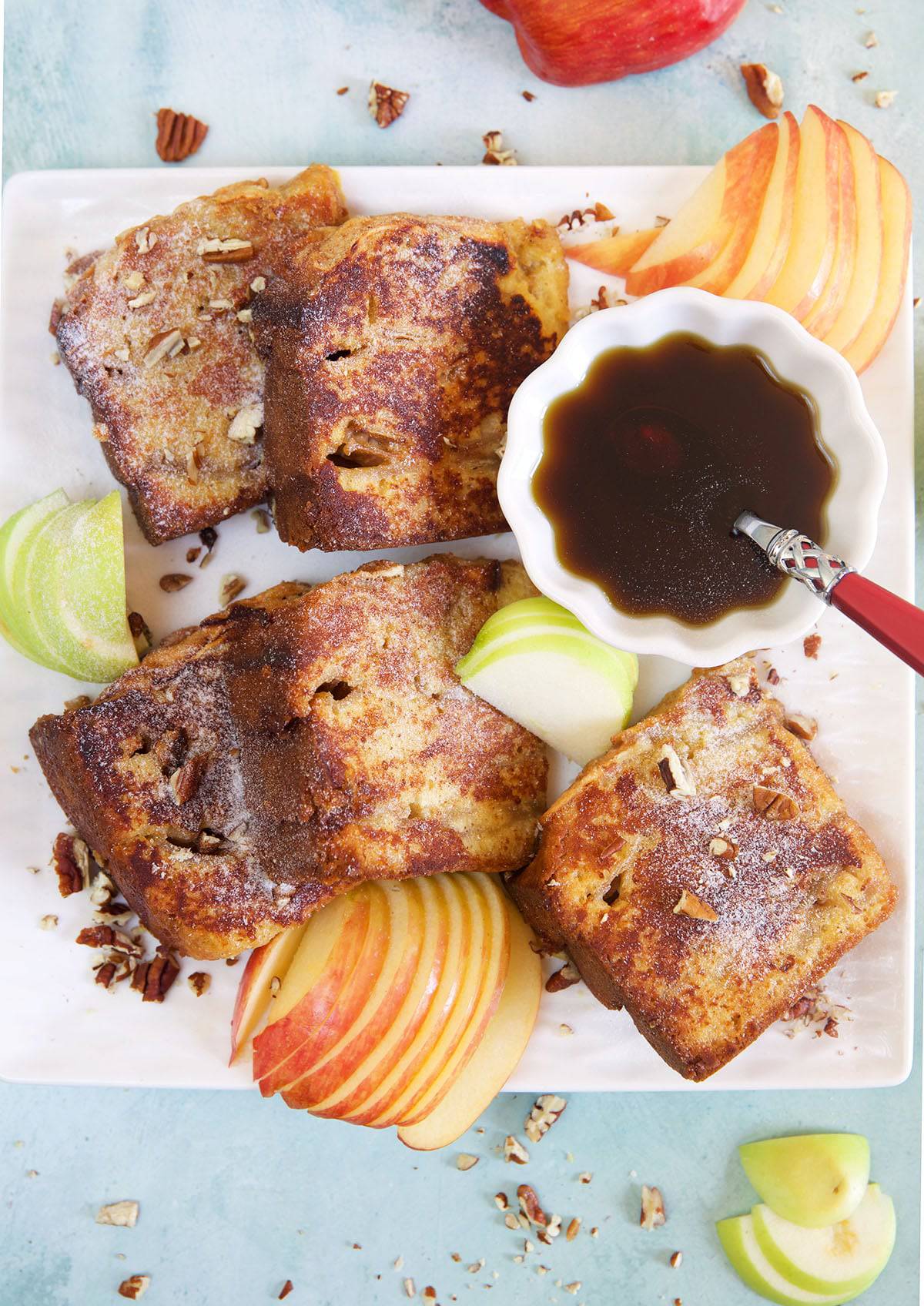 Several slices of French toast are placed on a white serving platted next to pieces of sliced apples. 