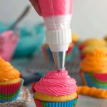 A piping bag is piping pink frosting onto a vanilla cupcake.